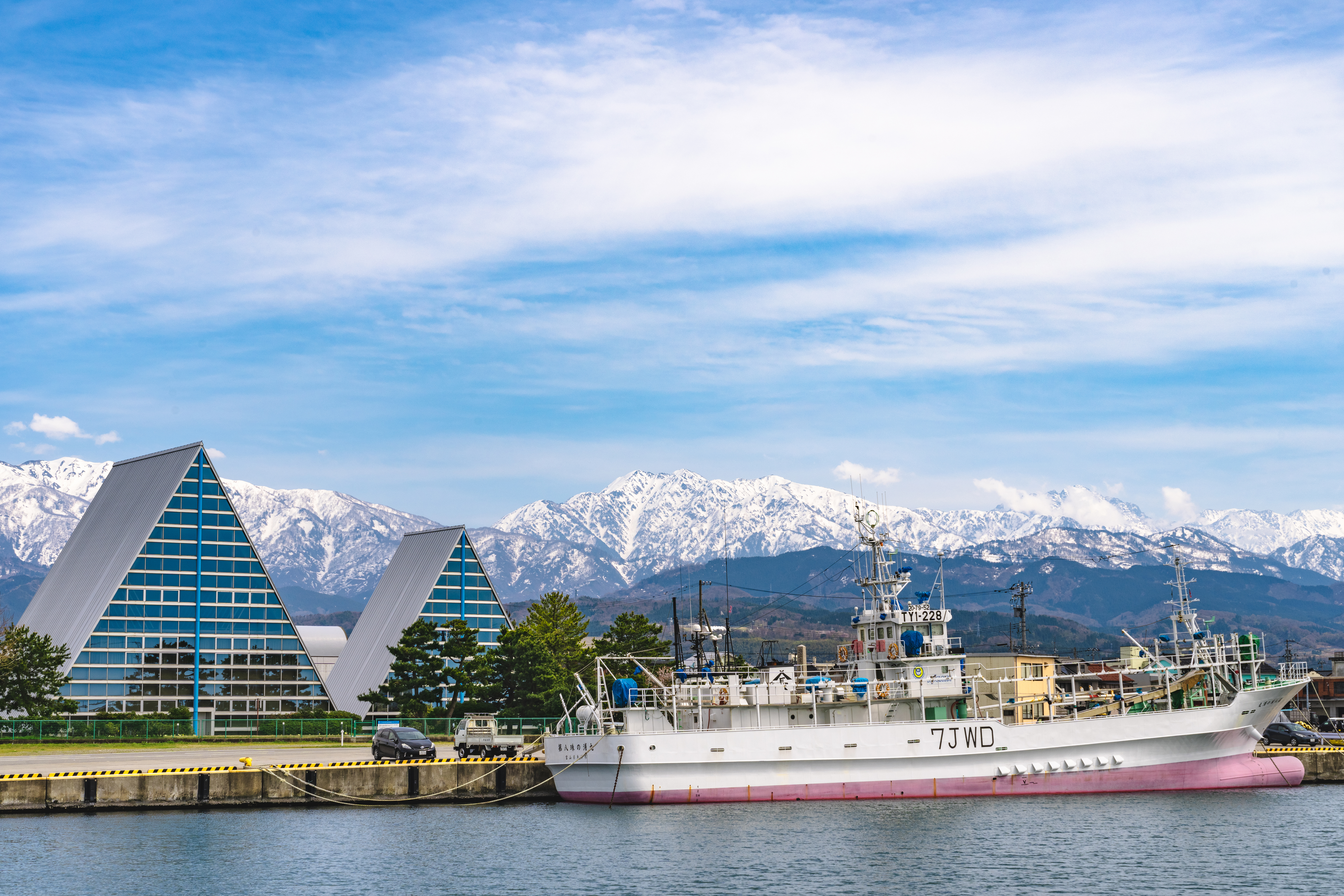 富山県魚津市の魚津漁港と立山連峰。三角形の特徴的な建物(魚津埋没林博物館)と雪をかぶった山々を背景に漁船が停泊している